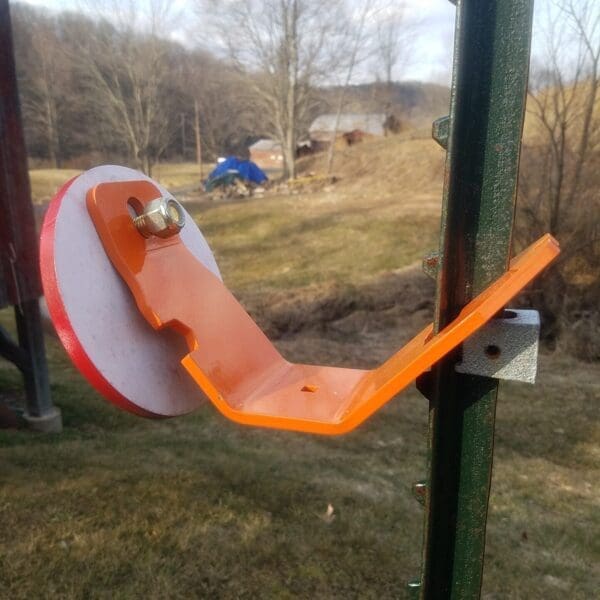 Orange L shaped metal mounting bracket with a round red wheel bolted to a green vertical post outdoors in a yard with trees in the background