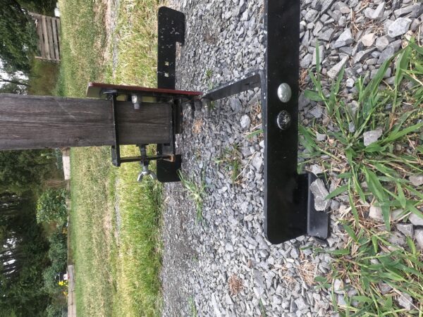 Black metal trailer hitch frame resting on a gravel surface beside a wooden postbeam assembly